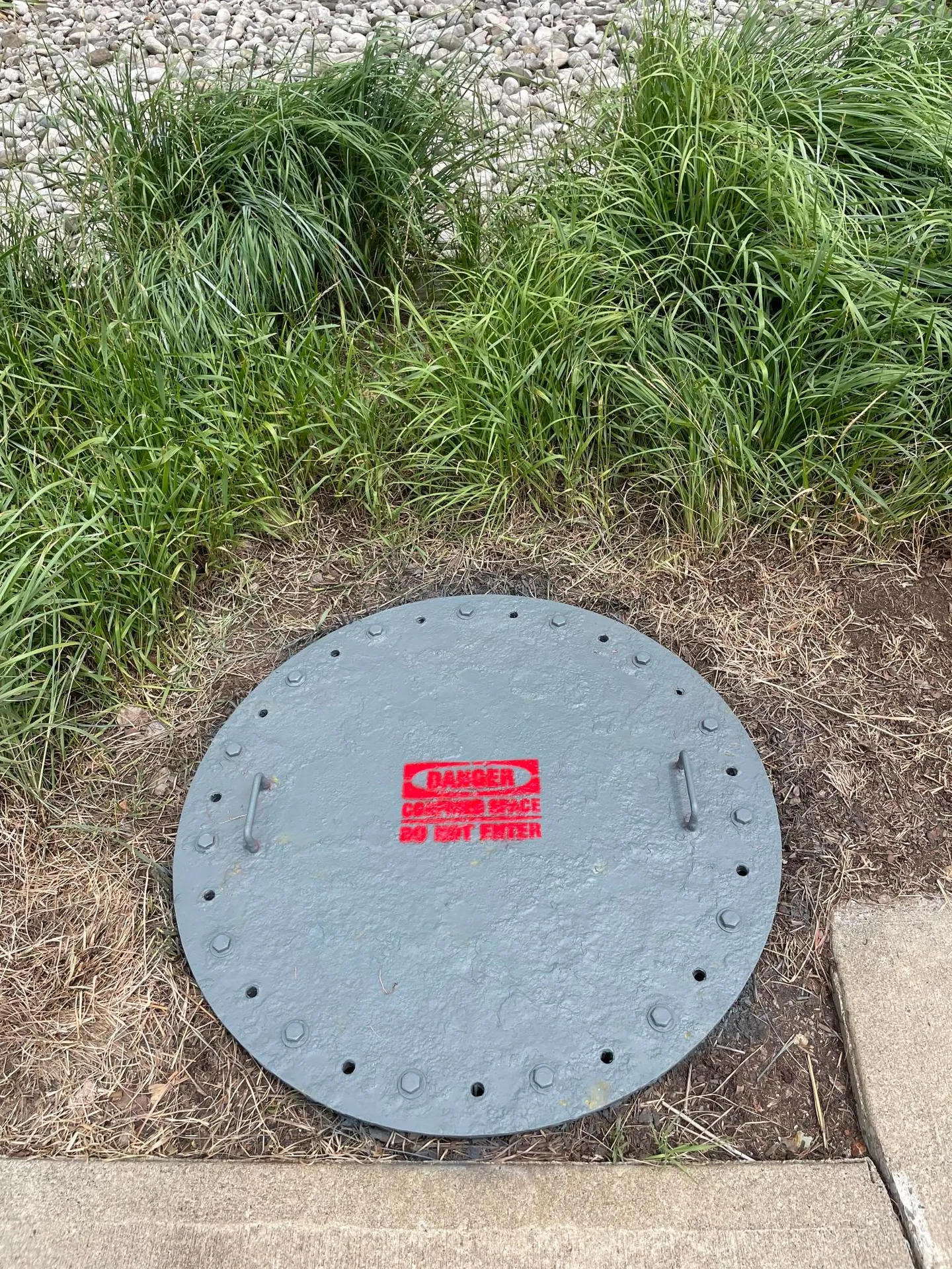 Freshly refinished grey manhole cover with red safety stencil in a landscaped bed