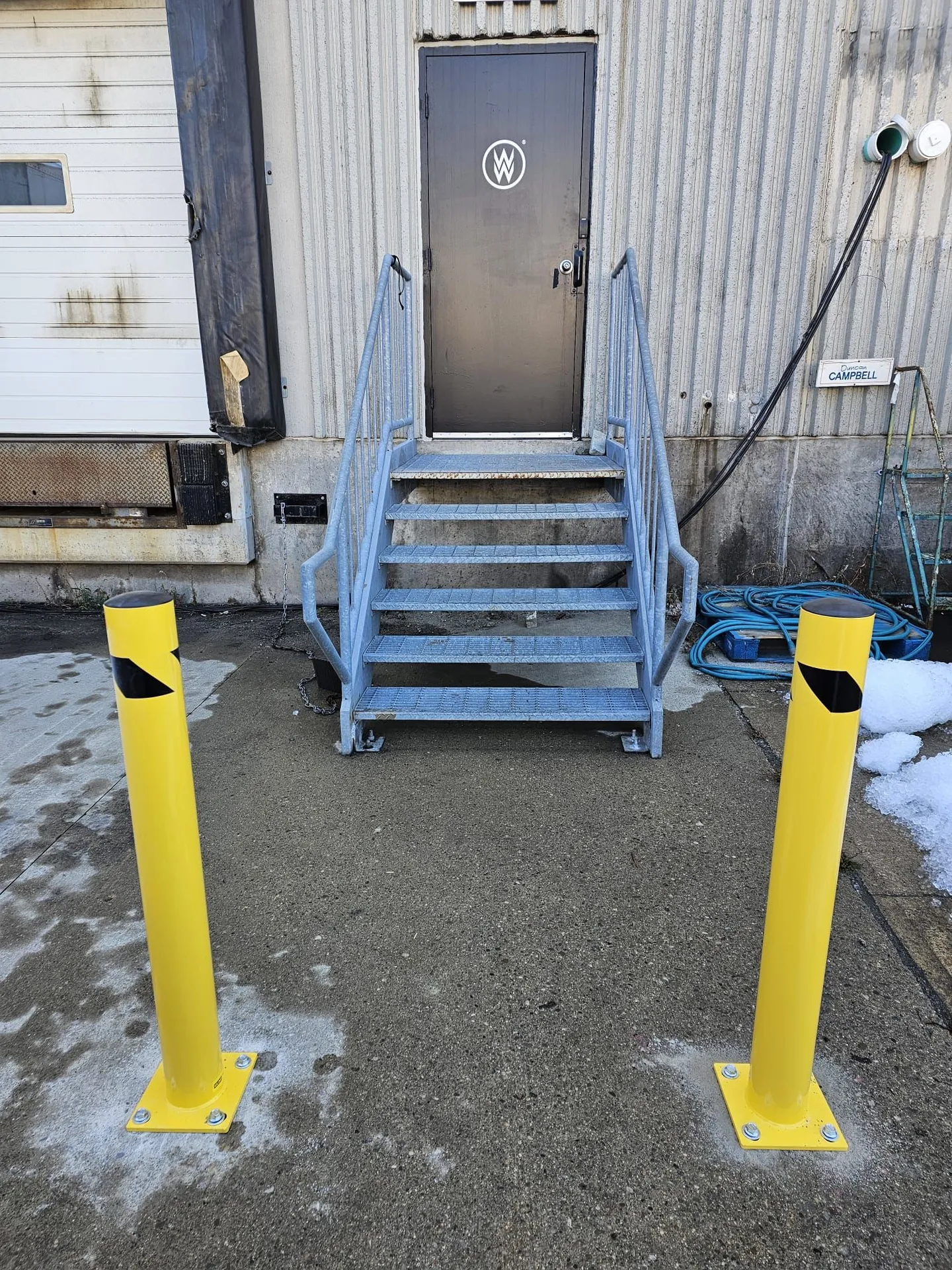 Two new yellow safety bollards installed at a loading-dock man-door beside a galvanized stair