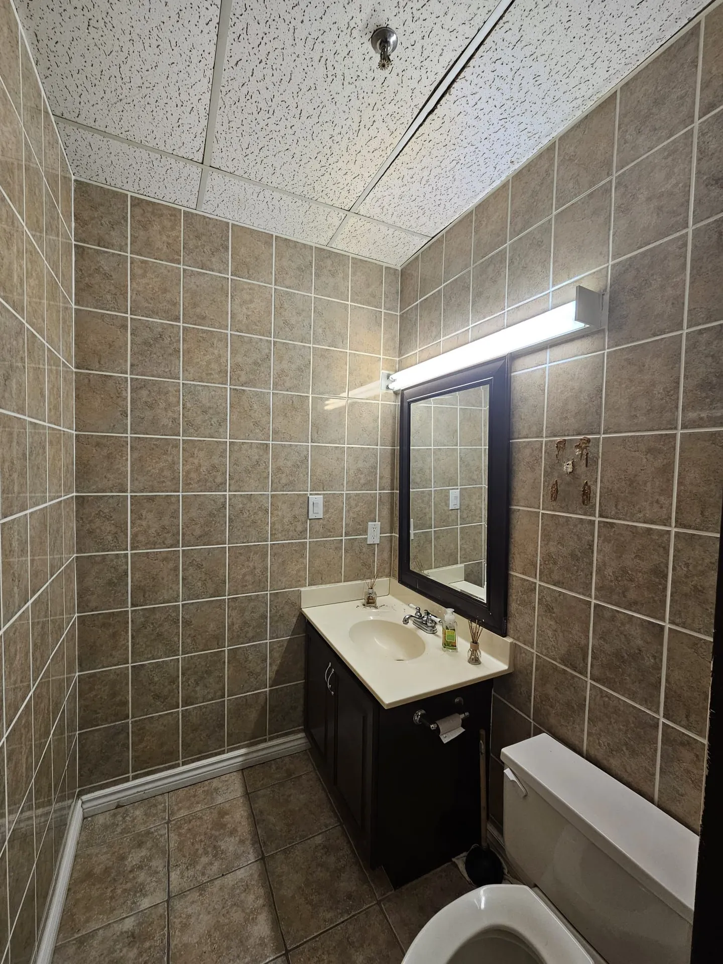 Dated washroom before renovation with floor-to-ceiling beige tile and old vanity and toilet
