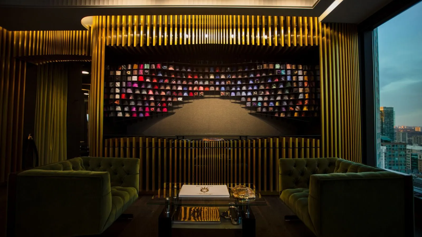 Fabric-swatch feature wall framed by gold slats with green velvet lounge chairs and a skyline window