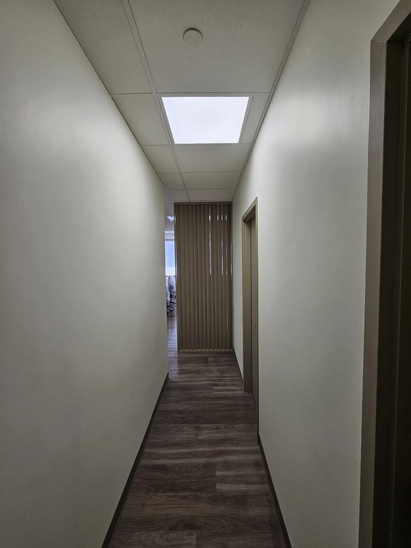 New narrow corridor with LVP flooring, crisp white walls, and vertical blinds at the end